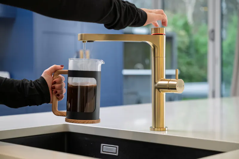 Person filling a French press with hot water from a brushed gold kitchen faucet
