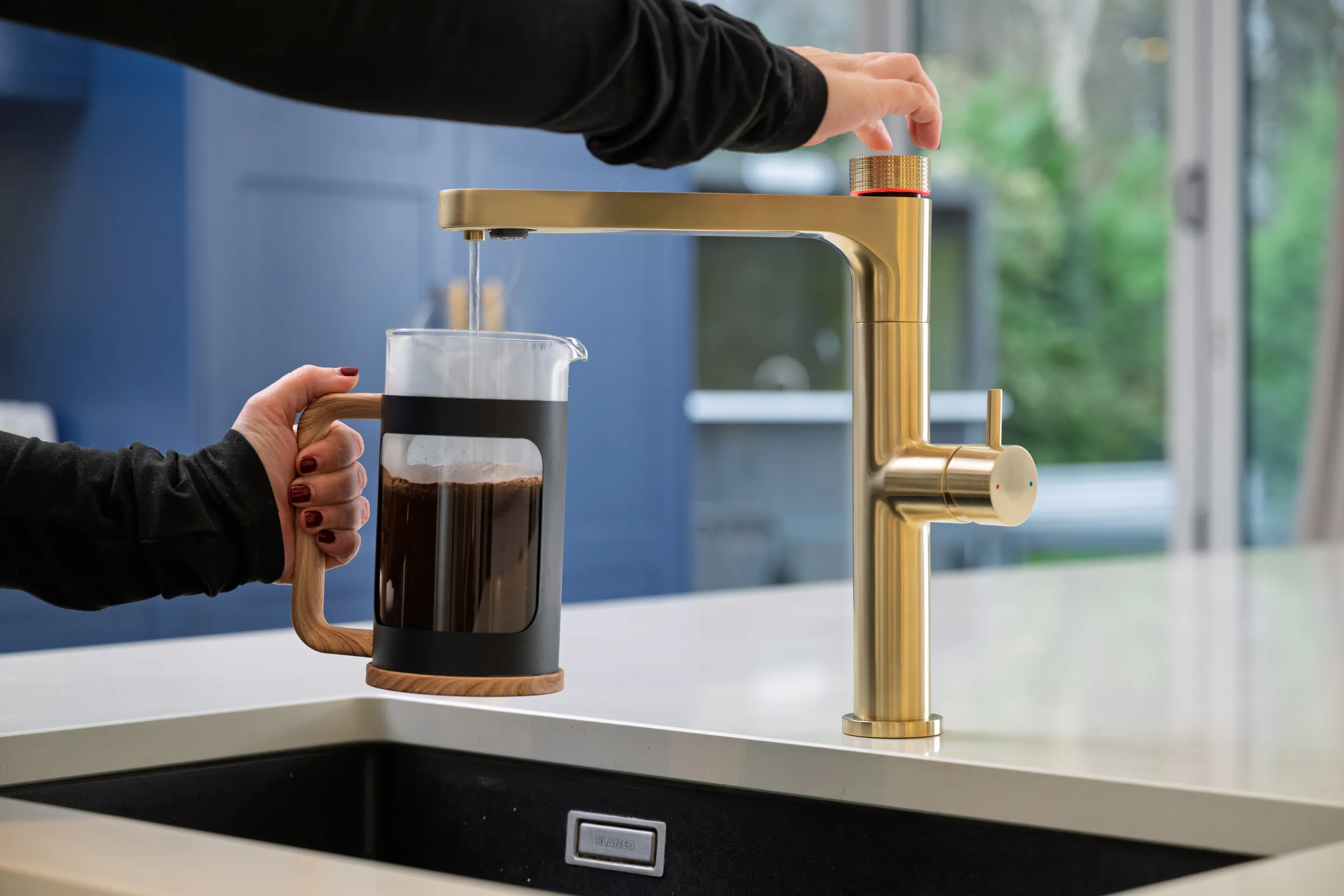 Person filling a French press with hot water from a brushed gold kitchen faucet