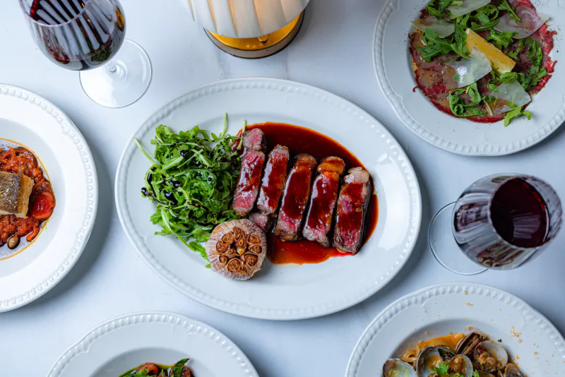 Sliced duck breast with red wine sauce, roasted garlic, and arugula on a white plate, surrounded by wine glasses and other gourmet dishes on a white table