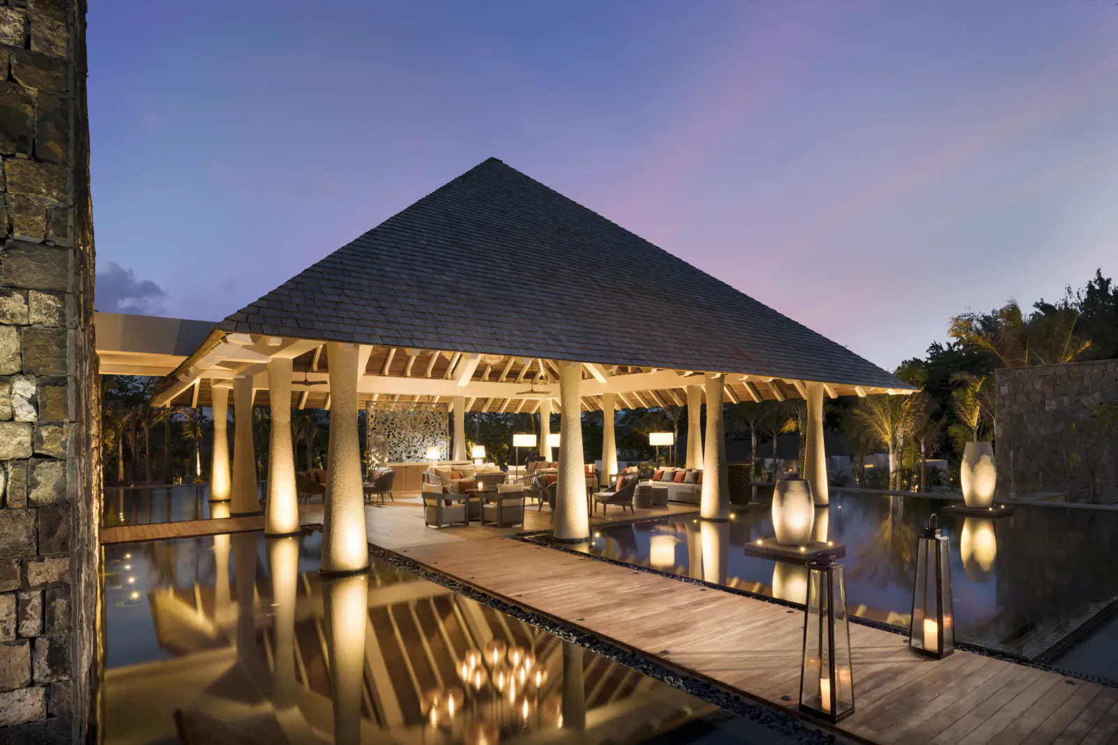 Luxury open-air lounge at dusk with thatched roof, wooden deck over reflecting pool, and glowing lanterns at Constance Le Chaland, Mauritius