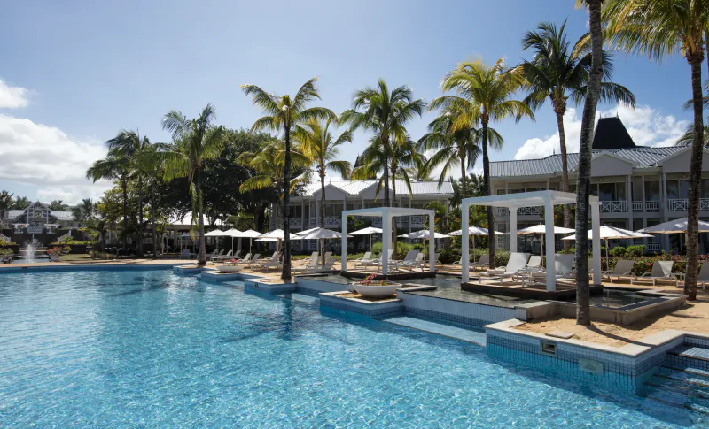 Luxury resort pool with palm trees, white umbrellas, and tropical buildings under blue sky