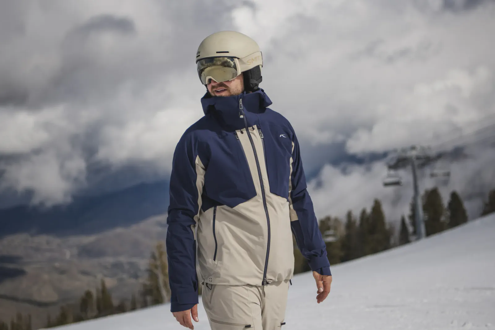 Man in blue and beige ski jacket and helmet standing on snowy slope with ski lift and mountains behind
