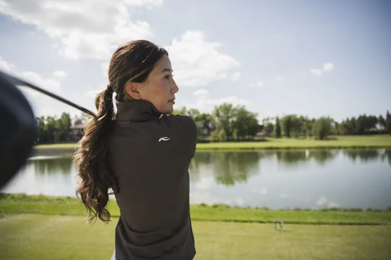 Woman in black golf jacket mid-swing on a sunny course with lake and trees behind