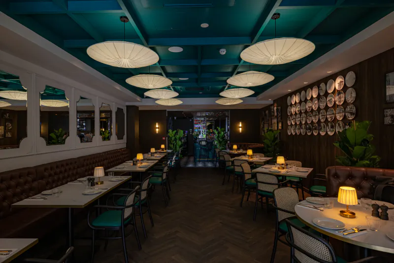 Elegant restaurant interior with teal ceiling, white pendant lights, dark wood walls with decorative plates, and neatly set dining tables with green chairs