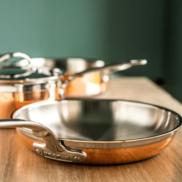 Close-up of a Hestan stainless steel and copper frying pan on a wooden surface with other cookware in the background