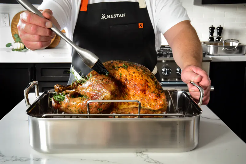 Chef basting roasted turkey with herbs in stainless roasting pan on kitchen counter