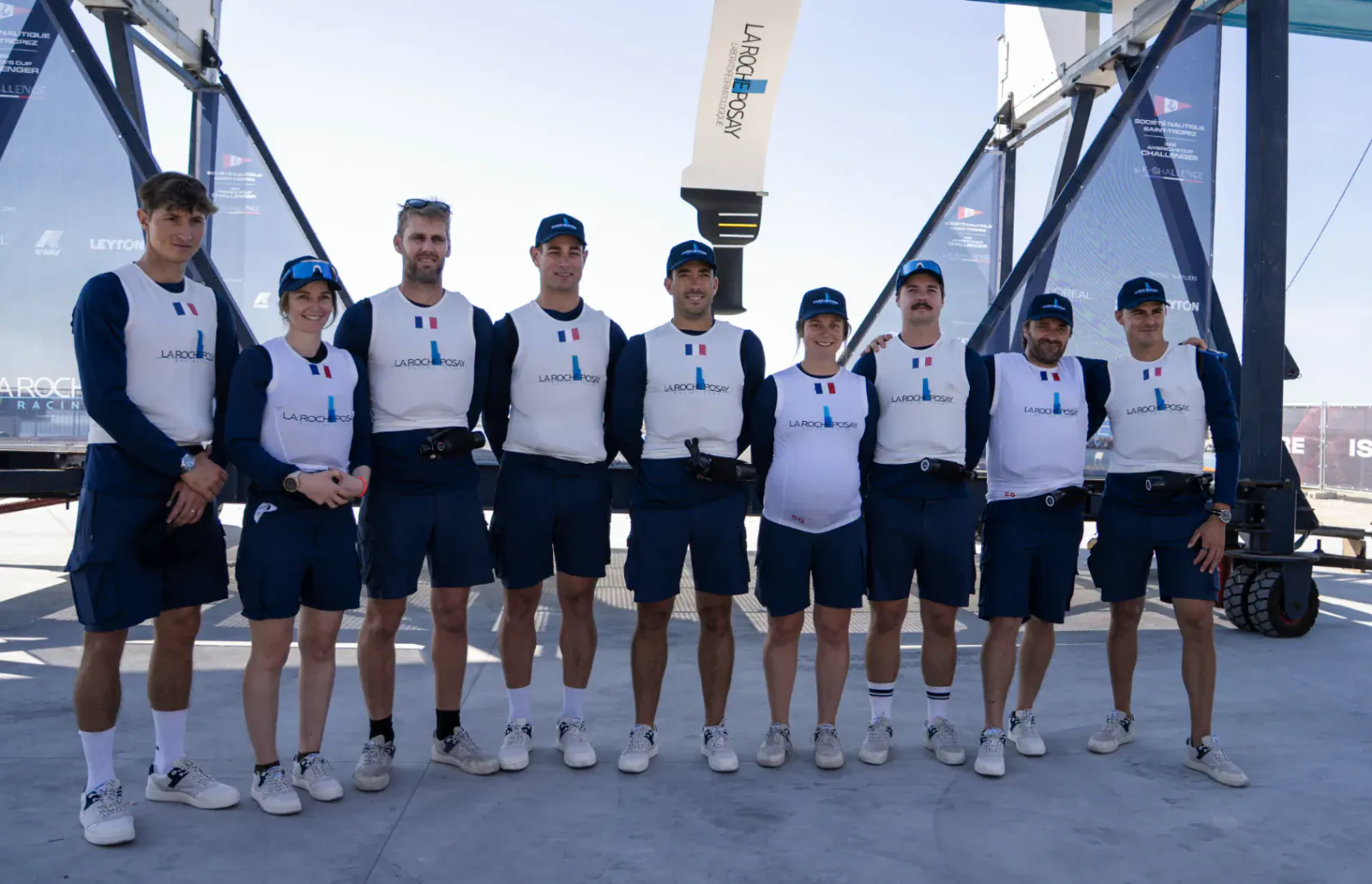 La Roche-Posay sailing team in matching uniforms posing together on a dock with AC75 boat and sponsor banners behind them