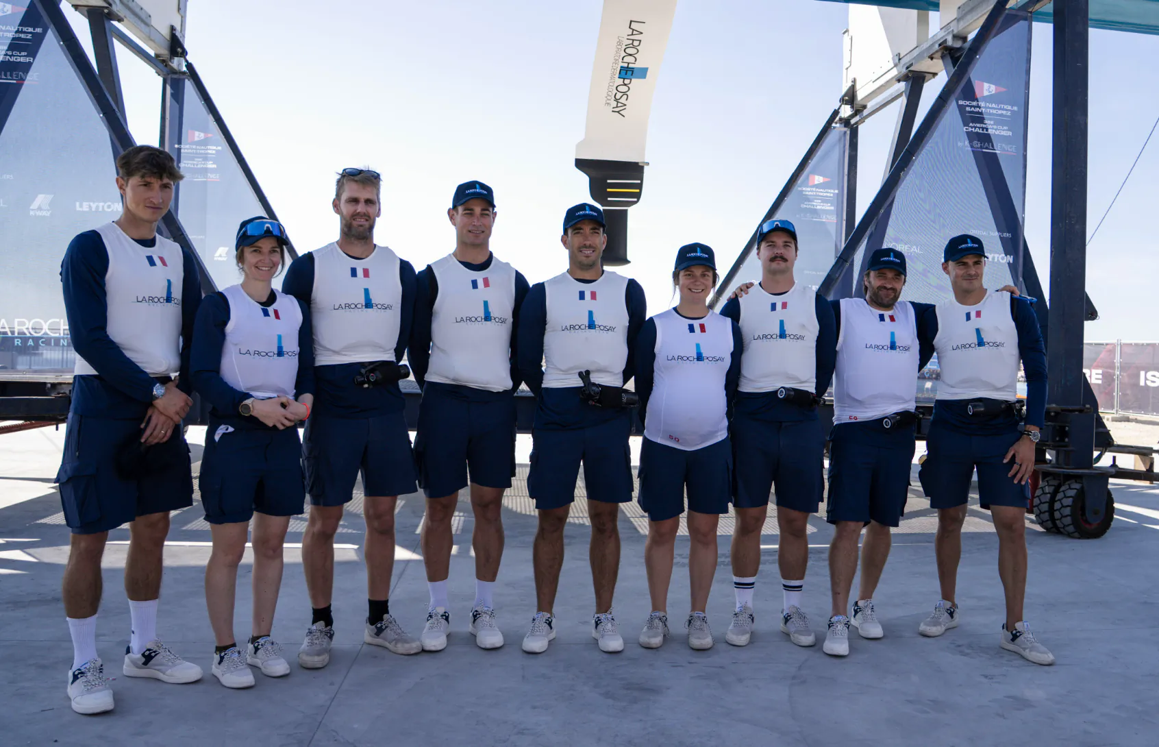 La Roche-Posay sailing team in matching uniforms posing together on a dock with AC75 boat and sponsor banners behind them