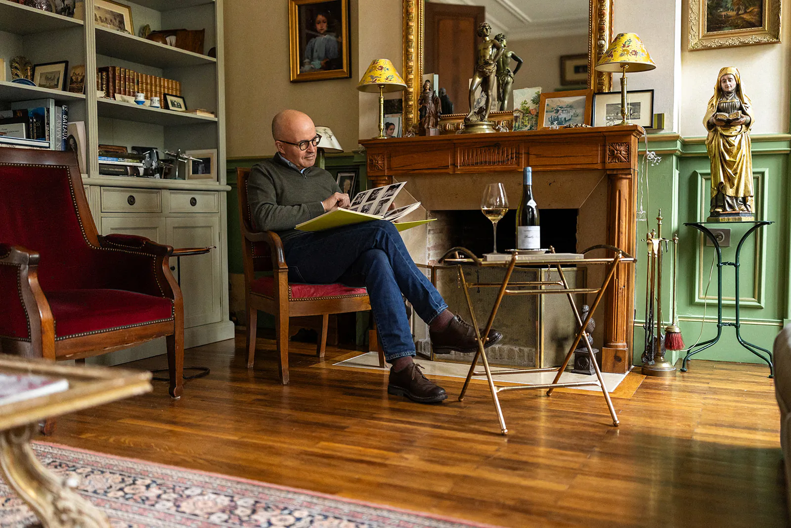 Bald man in glasses reading a magazine by a fireplace in a classic living room with wine glass and bottle on side table