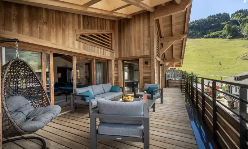 Modern wooden chalet balcony with grey sofa, hanging egg chair, and mountain valley view