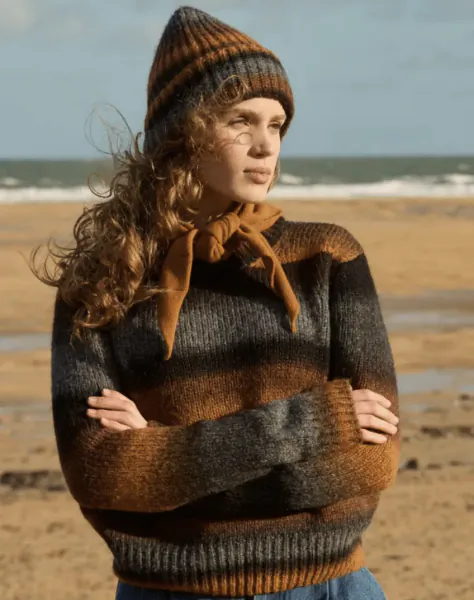 Woman with curly hair wearing striped knit sweater, beanie and scarf stands with arms crossed on a windy beach