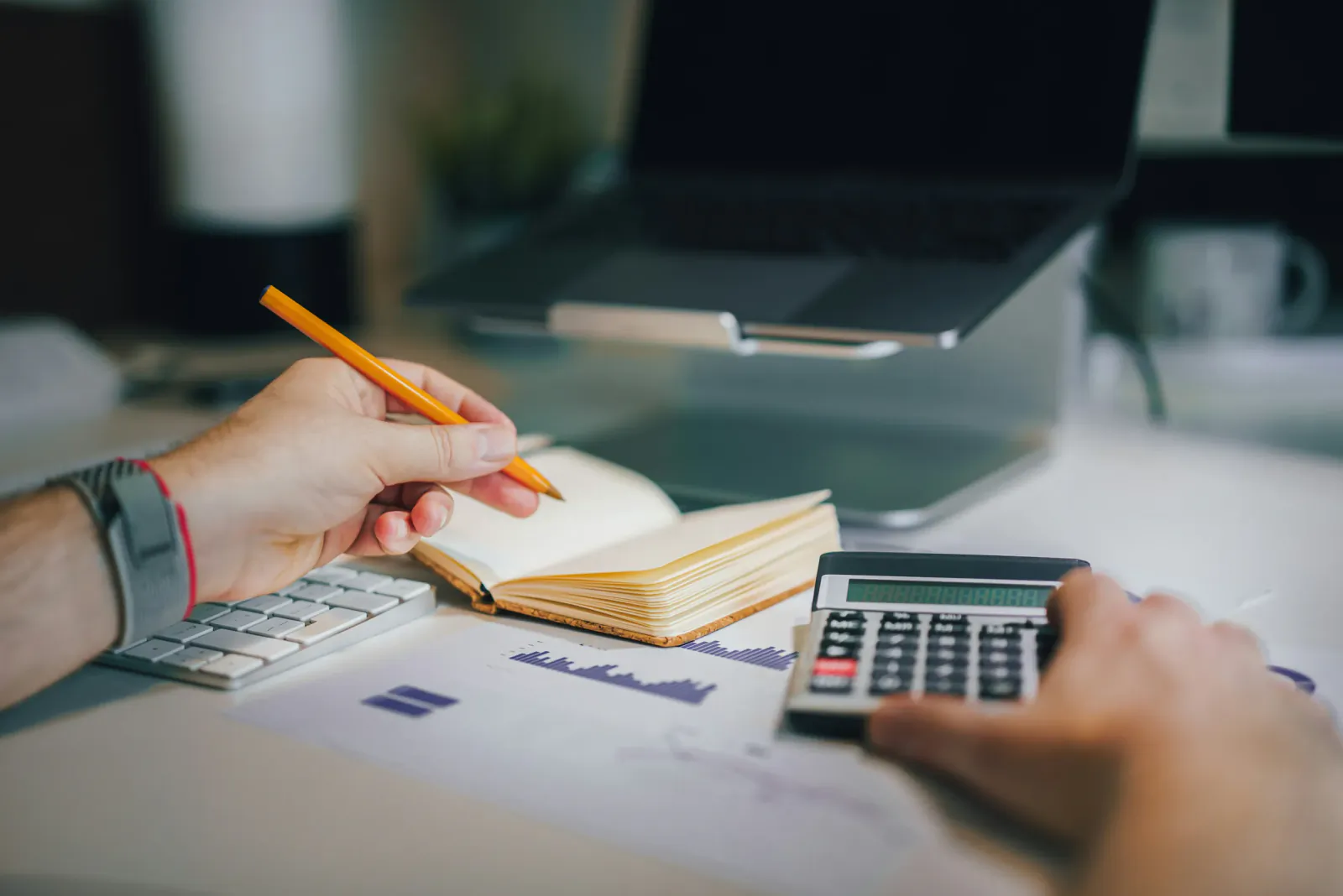 Hands using a calculator and writing in a notebook on a desk with financial charts and a laptop