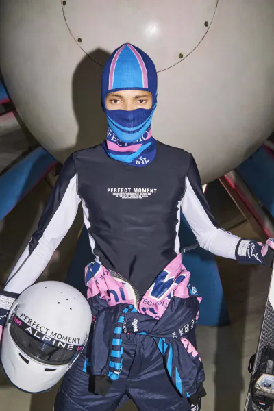 Racer in blue-pink balaclava and Perfect Moment Alpine suit holding white helmet with sponsor logos