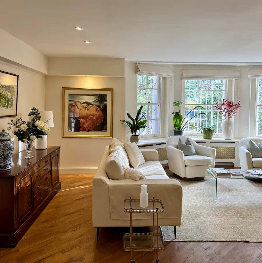 Bright living room with cream sofa, wooden sideboard, large floral painting, and three windows overlooking greenery