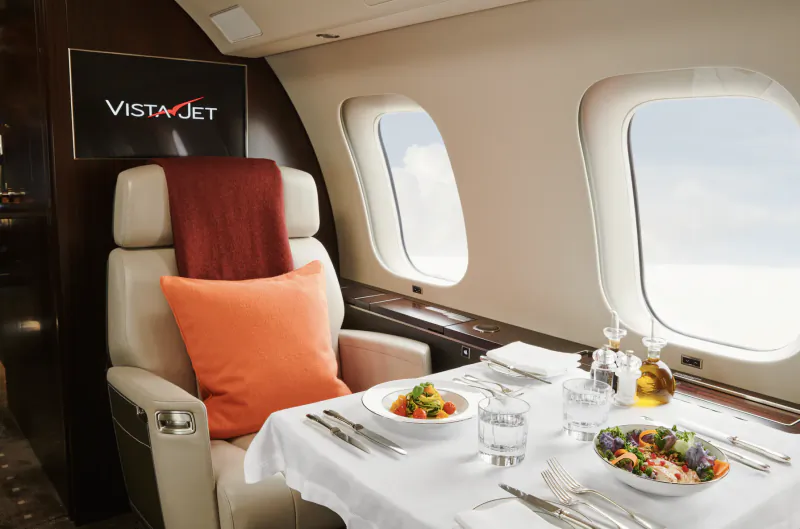 VistaJet private jet cabin with beige leather seat, orange pillow, red blanket, and two plated meals on white tablecloth beside large windows