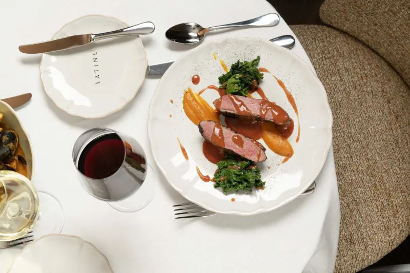 Plated sliced steak with green vegetables and orange sauce on white plate beside Latine branded dish, wine glass and cutlery on white tablecloth