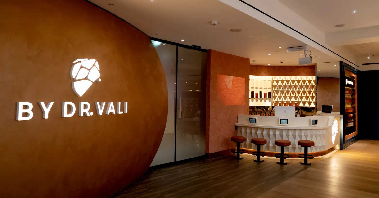 Modern reception area with illuminated 'BY DR. VALI' logo on curved terracotta wall and white curved desk with stools