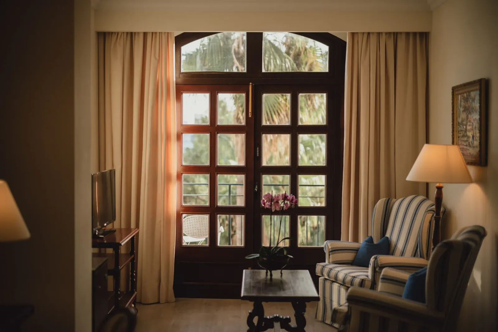 Cozy hotel lounge with striped armchairs, a vase of pink flowers on a coffee table, and large wooden doors opening to palm trees outside