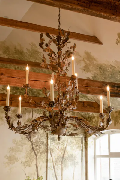 Ornate metal chandelier with lit candles hanging from wooden beams against a forest mural wall