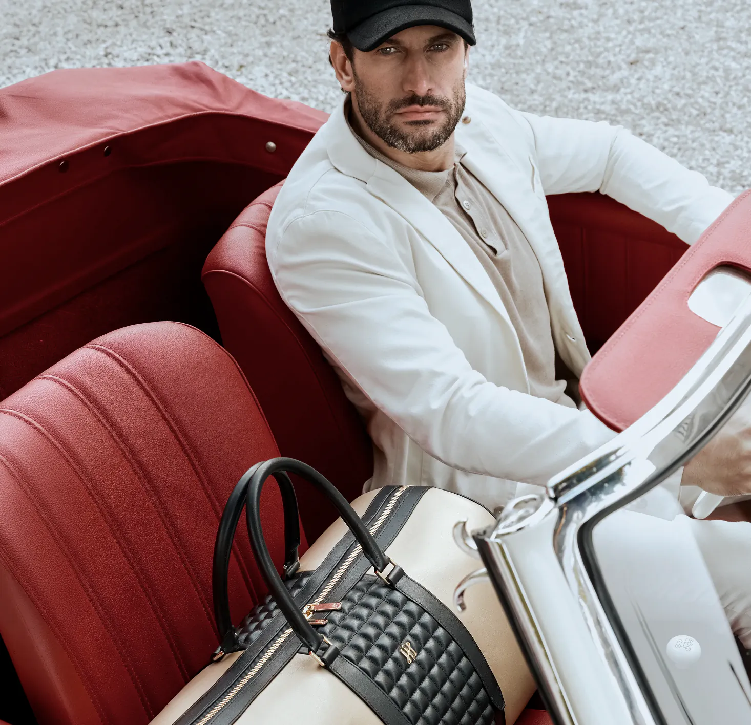 Man in white blazer and black cap seated in red vintage convertible with luxury duffel bag on seat