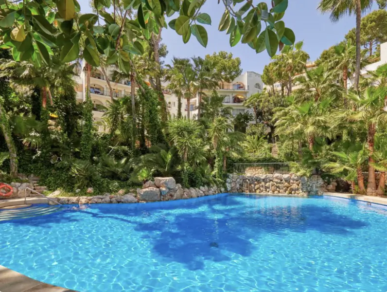 Resort swimming pool surrounded by palm trees and lush greenery with multi-story hotel buildings in the background