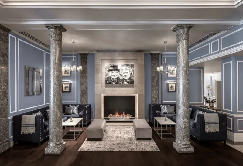 Elegant hotel lounge with two marble columns, blue velvet sofas, central fireplace, and black-and-white wall art