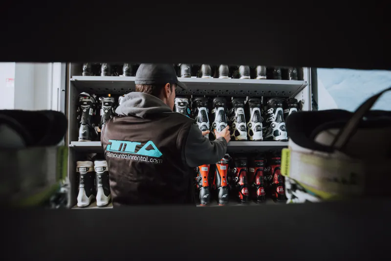 Technician in All Mountain Rental vest organizing ski boots on wall shelves in a rental shop