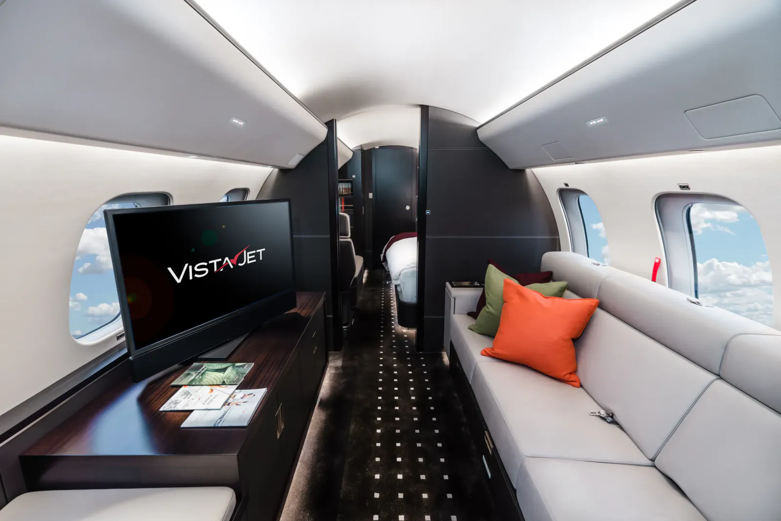 VistaJet private jet cabin interior with white leather seating, orange and green pillows, wood desk, and VistaJet logo on TV screen
