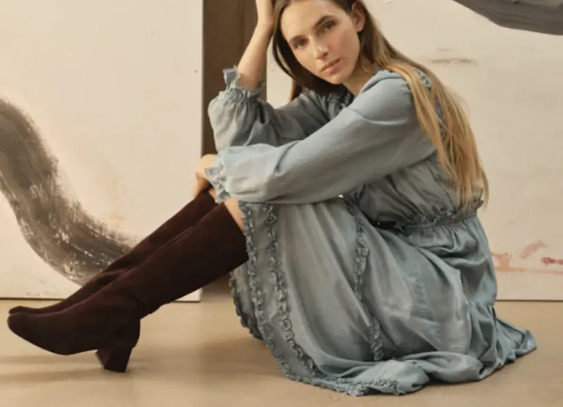 Woman in light blue dress and brown suede boots sitting on floor with hand in hair
