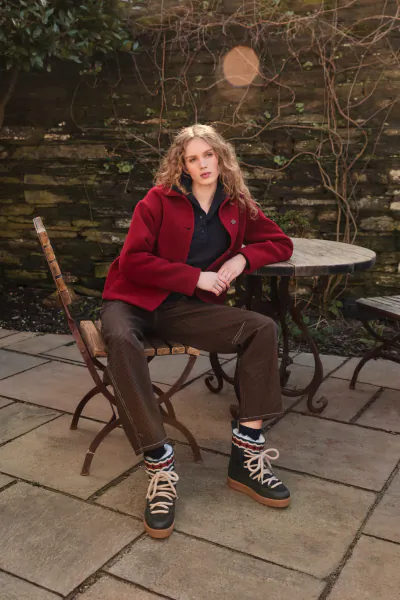 Woman with long wavy blonde hair wearing a red fleece jacket and brown trousers sits on a wooden chair at a stone patio table, wearing dark green lace-up boots