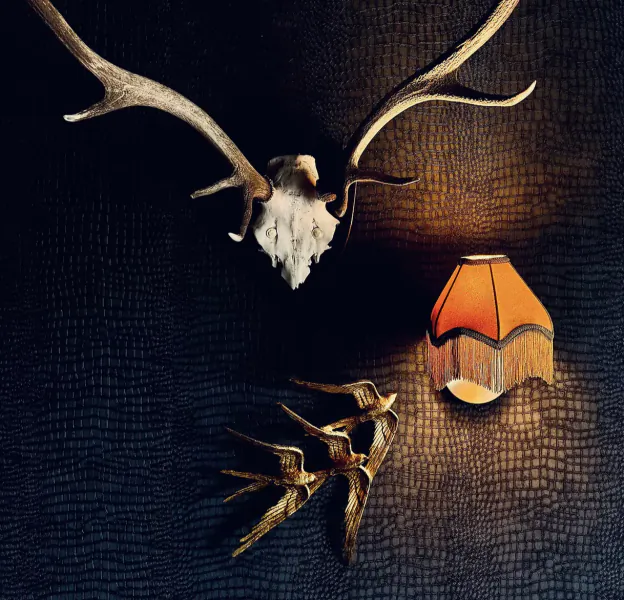 Mounted deer skull with antlers, golden swallow bird ornament, and small fringed lampshade on dark textured leather background