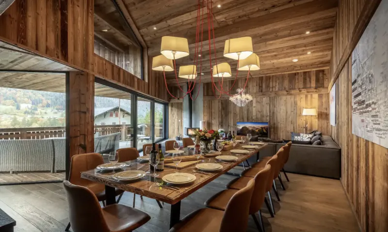 Modern alpine dining room with long wooden table set for dinner, brown leather chairs, red pendant lights, and mountain view windows