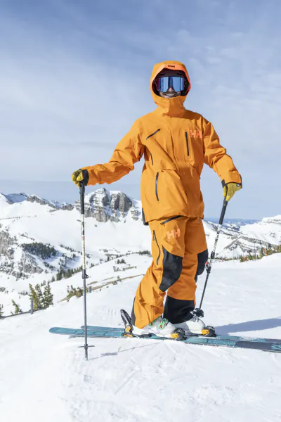 Skier in bright orange Helly Hansen snowsuit and goggles standing on skis with poles on snowy mountain slope