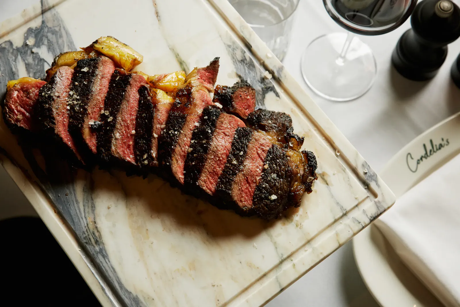 Sliced medium-rare steak with charred edges on marble board, wine glass and pepper mill nearby, Cordelia's logo visible