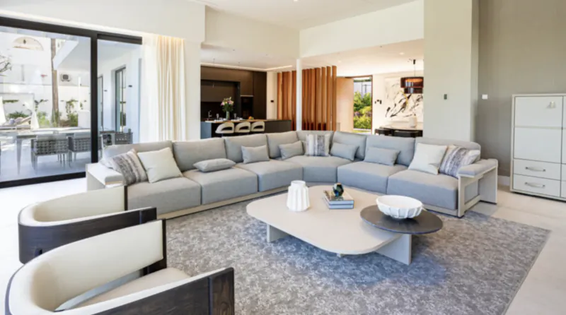 Modern open-plan living room with large grey sectional sofa, coffee table on grey rug, and open kitchen in luxury villa