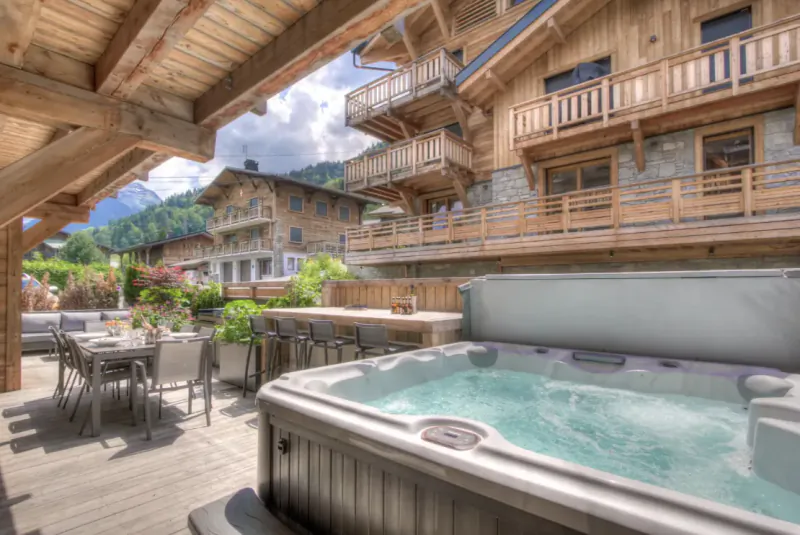 Wooden chalet deck with bubbling hot tub, outdoor dining table, and mountain views in Morzine