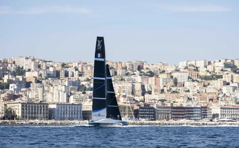 Black AC75 racing yacht with Louis Vuitton logo sailing past Naples waterfront cityscape