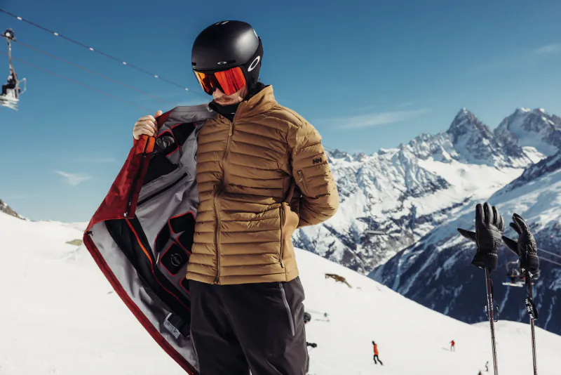 Skier in tan puffer jacket and black helmet opens jacket on snowy mountain slope with ski lift and peaks behind
