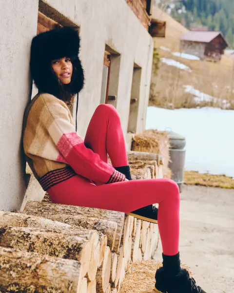 Woman in bright pink leggings and fur-trimmed hat sits on stacked logs outside a rustic building in snowy mountains