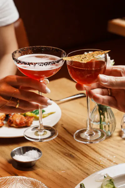 Two hands toasting with red cocktails in coupe glasses, one with black salt rim and the other garnished with a crisp, on a wooden restaurant table