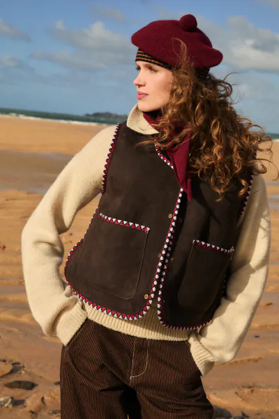 Woman in maroon beret and brown suede vest with colorful trim stands on a sandy beach, looking to the side