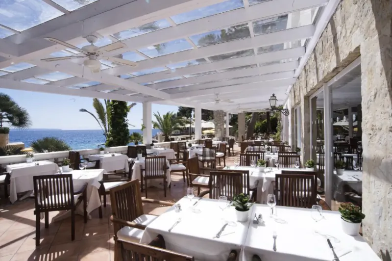 Elegant outdoor restaurant patio with white-clothed tables overlooking the sea under a glass-roofed pergola