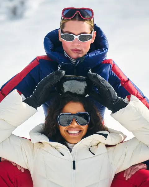 Two women in winter ski gear pose on snowy slope, one in white jacket smiling with goggles on, the other in blue-red jacket holding goggles above her head
