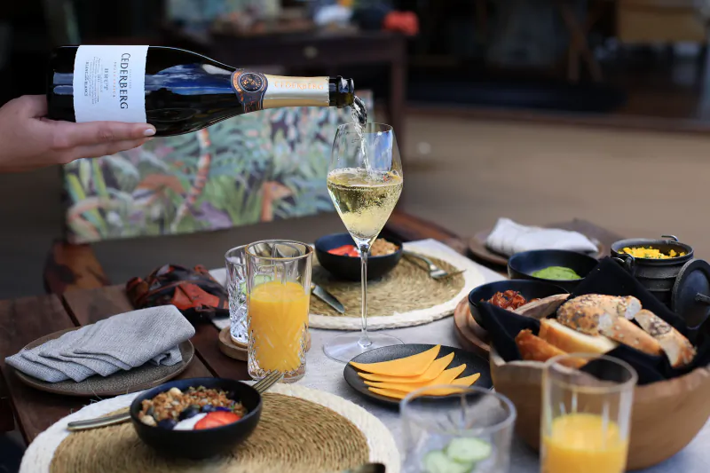Hand pouring G.H. Mumm champagne into a flute at a breakfast table with orange juice, bagels, cheese, and fruit bowls