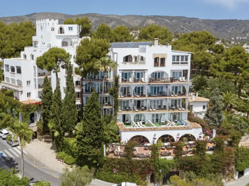 White multi-story Mediterranean hotel with balconies and pool, surrounded by trees and hills