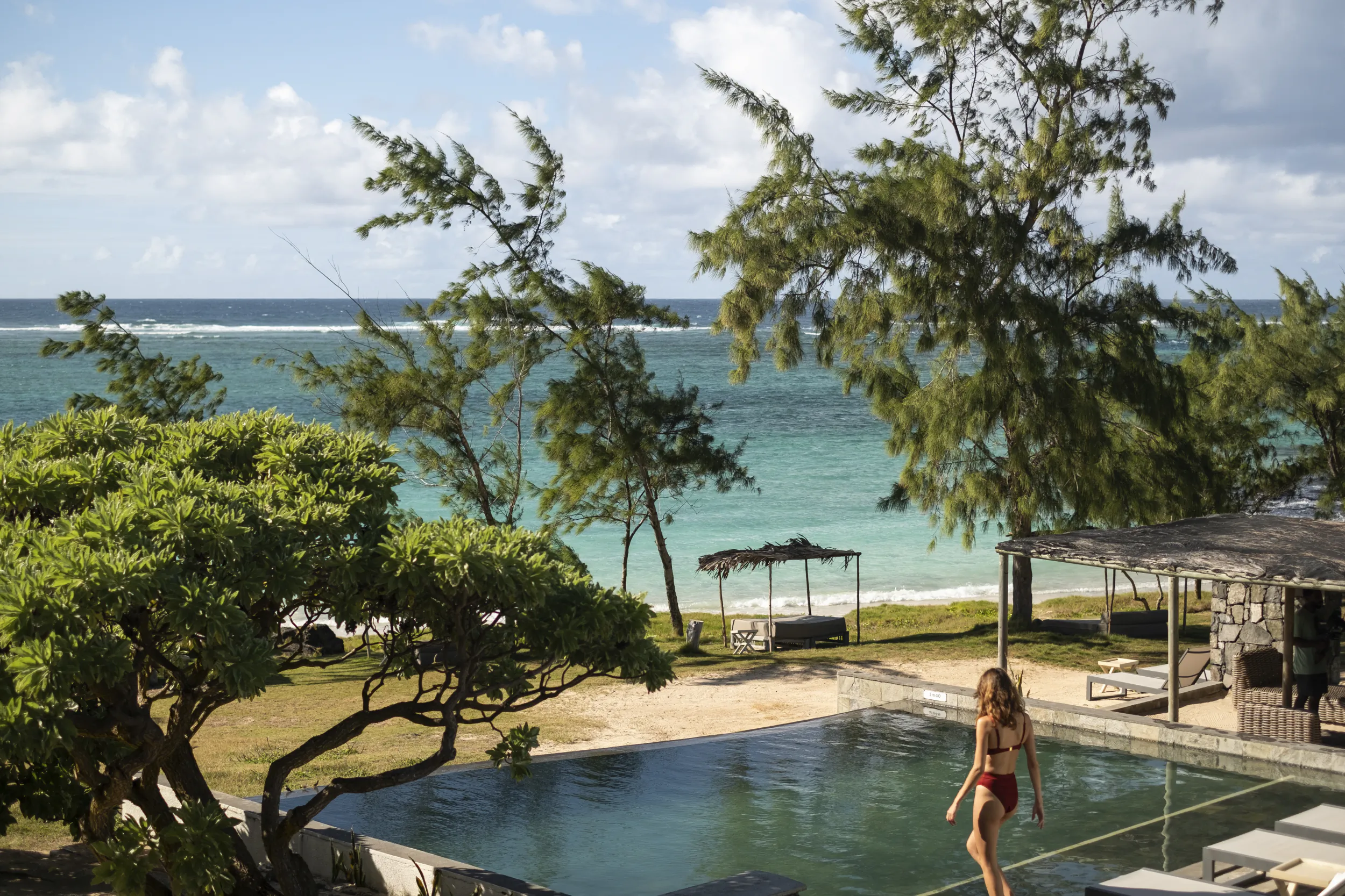 Woman in red swimsuit walking beside infinity pool overlooking turquoise ocean and tropical trees at Mauritius resort