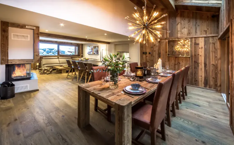 Rustic wooden dining table set with plates and flowers in a cozy alpine chalet with fireplace and modern chandelier