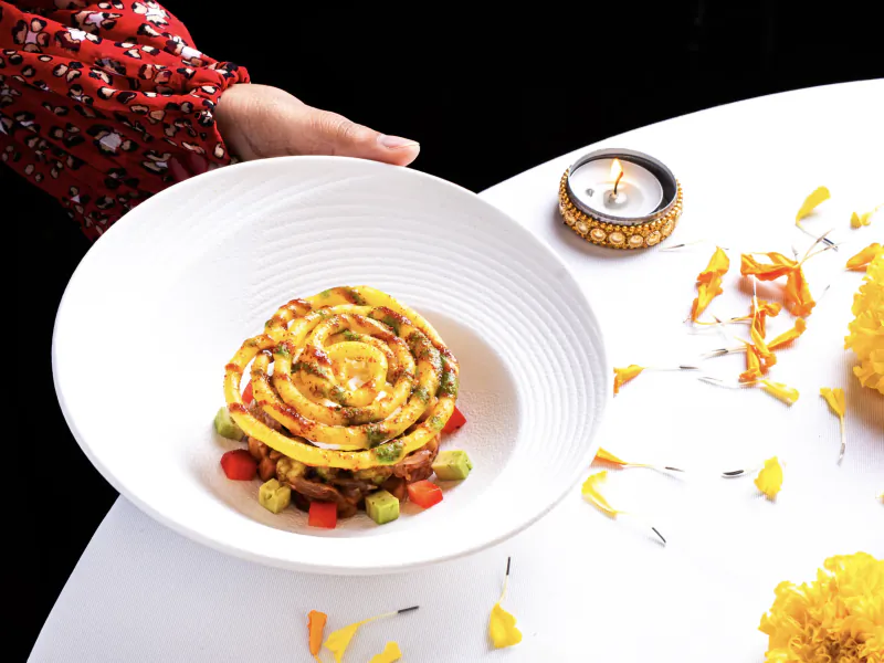 Hand presenting a white plate with a spiral yellow dish garnished with red sauce, diced vegetables, and edible flowers beside a lit candle on a white table