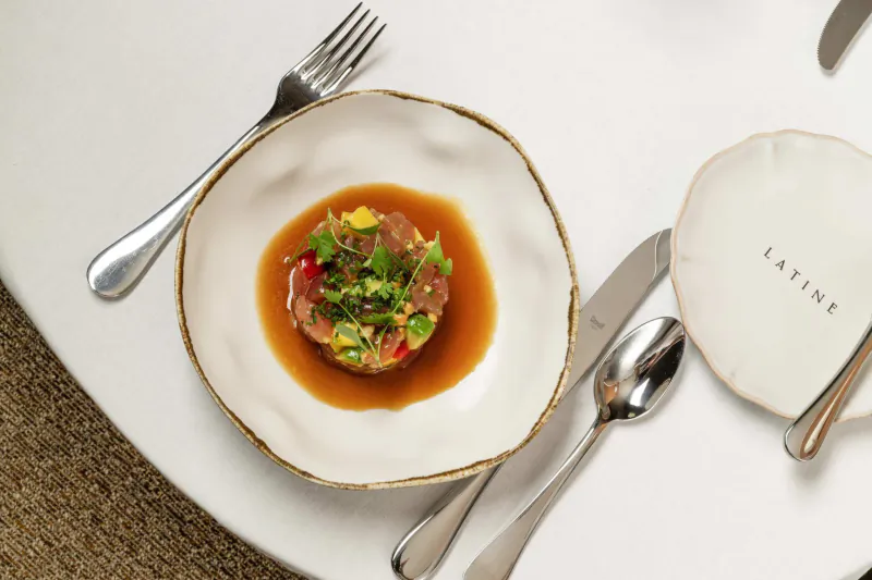 Tuna tartare with avocado and microgreens in brown sauce on white plate, silverware, and Latine branded plate on white table