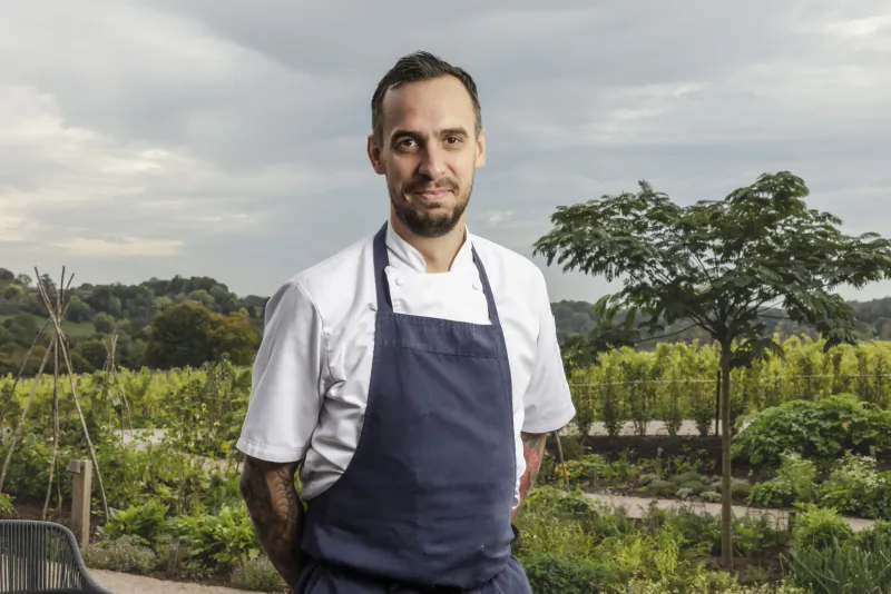 Male chef in white jacket and blue apron standing in vineyard garden with trees and hills